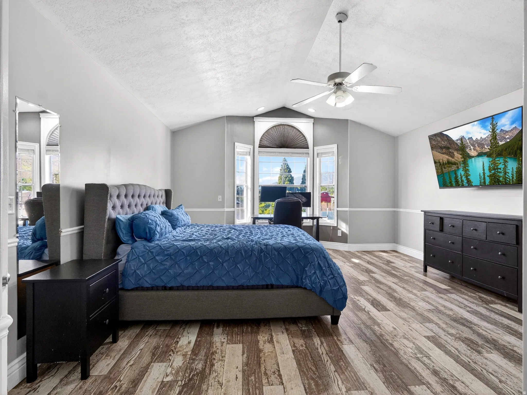 Spacious room featuring vaulted ceilings, wood-finish flooring, a ceiling fan, and multiple windows with a half-round transom