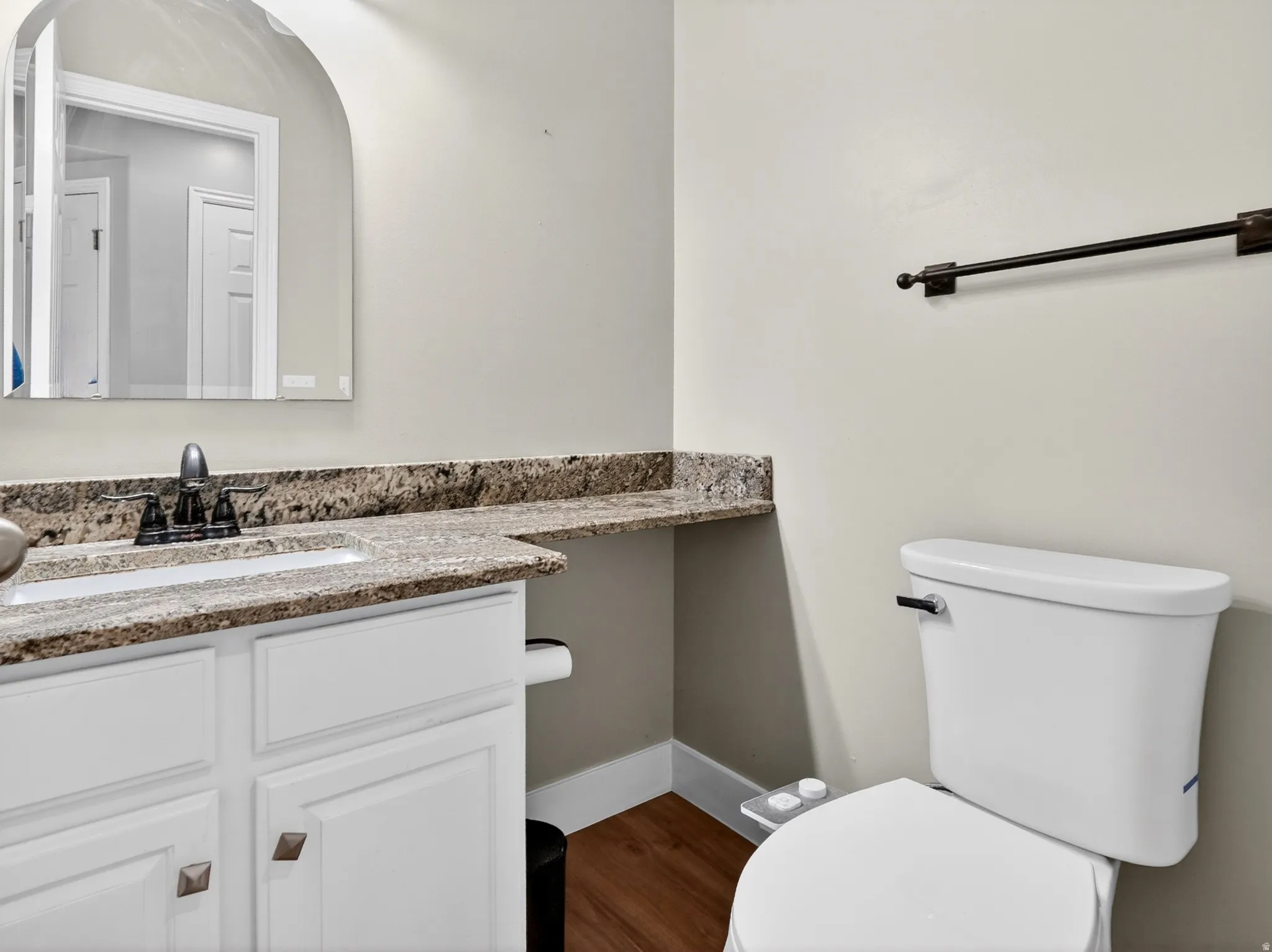 Bathroom featuring a white vanity with paneling and square bronze-finish hardware, a granite-finish countertop with an integrated sink, an arched mirror, a two-piece toilet, and wood-finish flooring