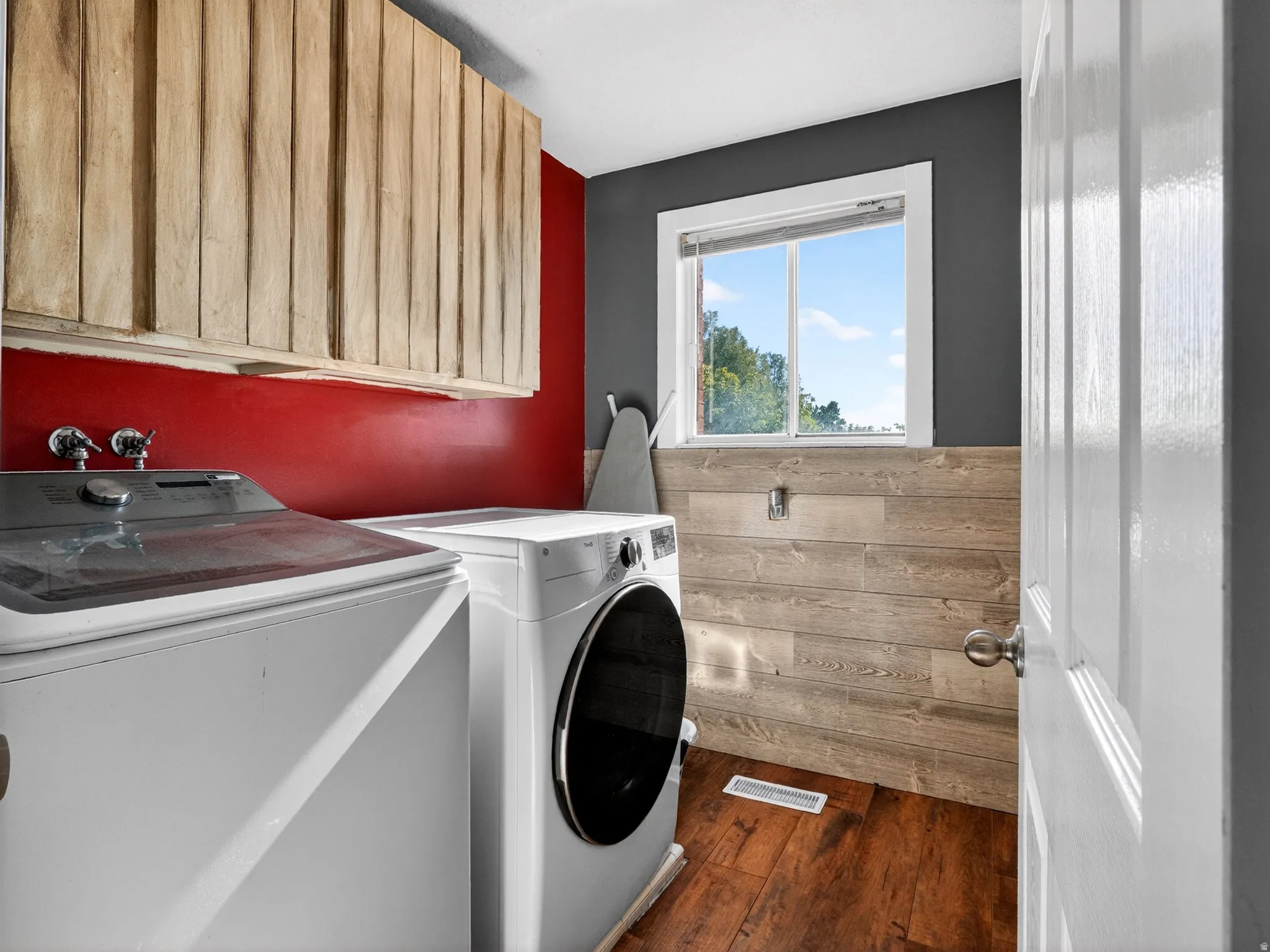 Utility space featuring wood-finish flooring, wood-panel accent wall, upper cabinetry with natural wood texture, and a white framed window