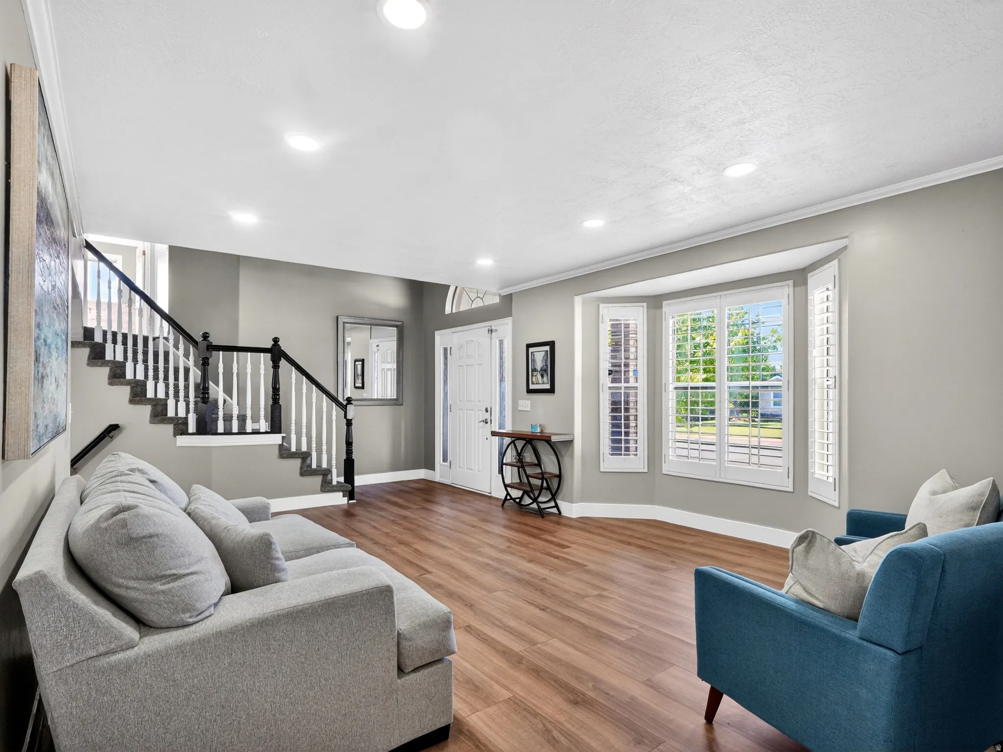 Spacious interior featuring wood-finish flooring, a prominent staircase with white risers and dark wood banister, recessed lighting, and a bay window with plantation shutters