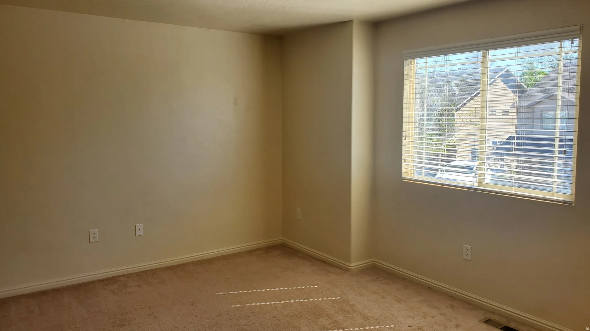 Spacious room with neutral wall tones, light brown carpeting, white baseboards, and a large window with white horizontal blinds