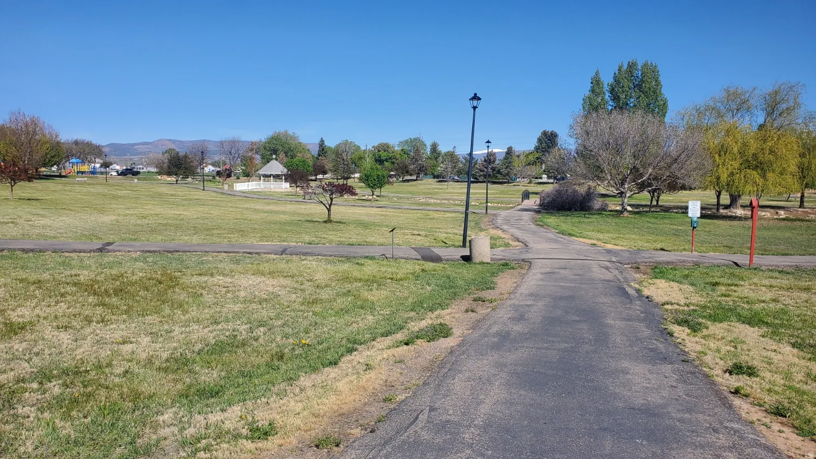 City park featuring paved walking paths, mature trees, and a distant gazebo