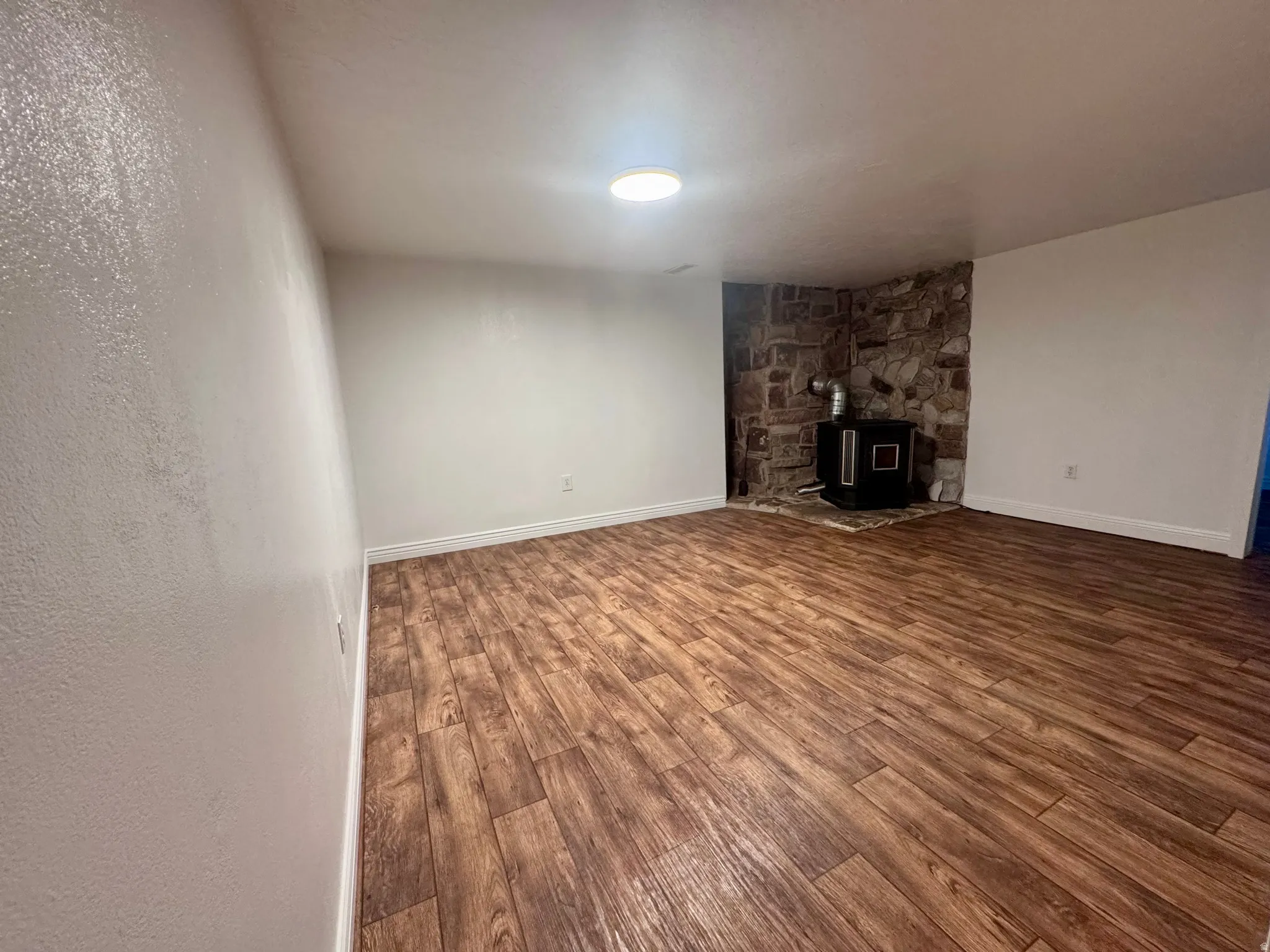 Spacious room featuring wood-finish flooring, white baseboards, and a stone accent wall with a black wood-burning stove