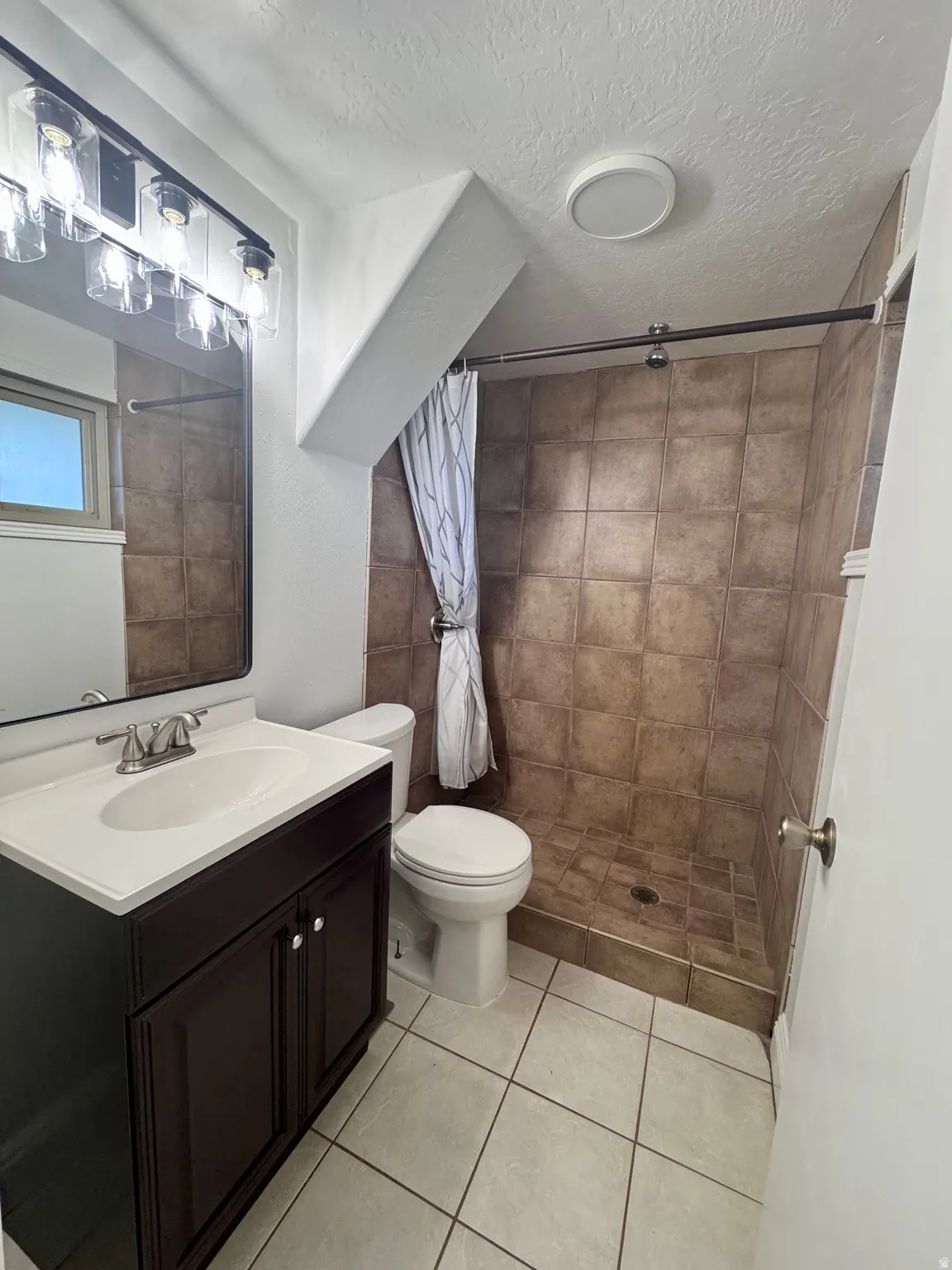 Bathroom featuring a dark wood-finish vanity with an integrated sink, a contemporary light fixture, a tiled shower enclosure, and white square floor tiles