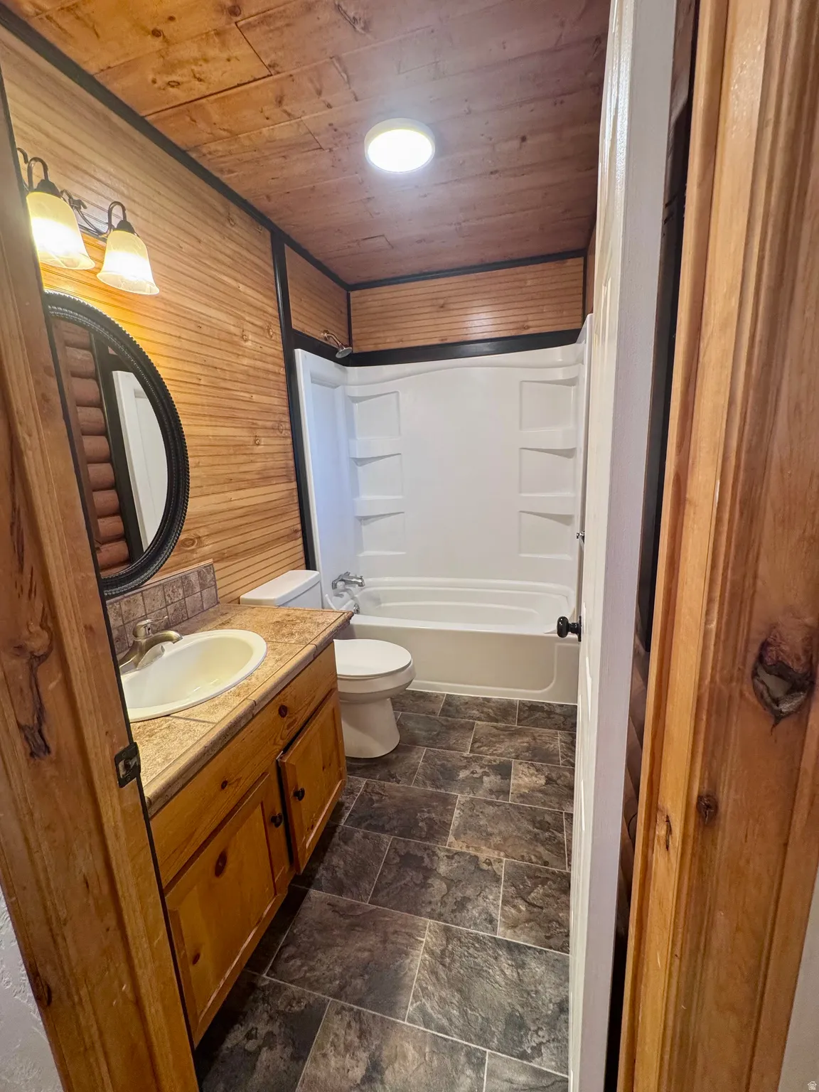 Bathroom featuring wood-finish wall and ceiling panels, a white tub/shower combination, a vanity with a light-toned countertop, and dark-toned tile flooring