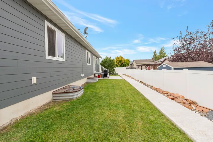 Side yard featuring a concrete walkway, manicured lawn, white vinyl fencing, and a dark gray siding exterior with white trim windows