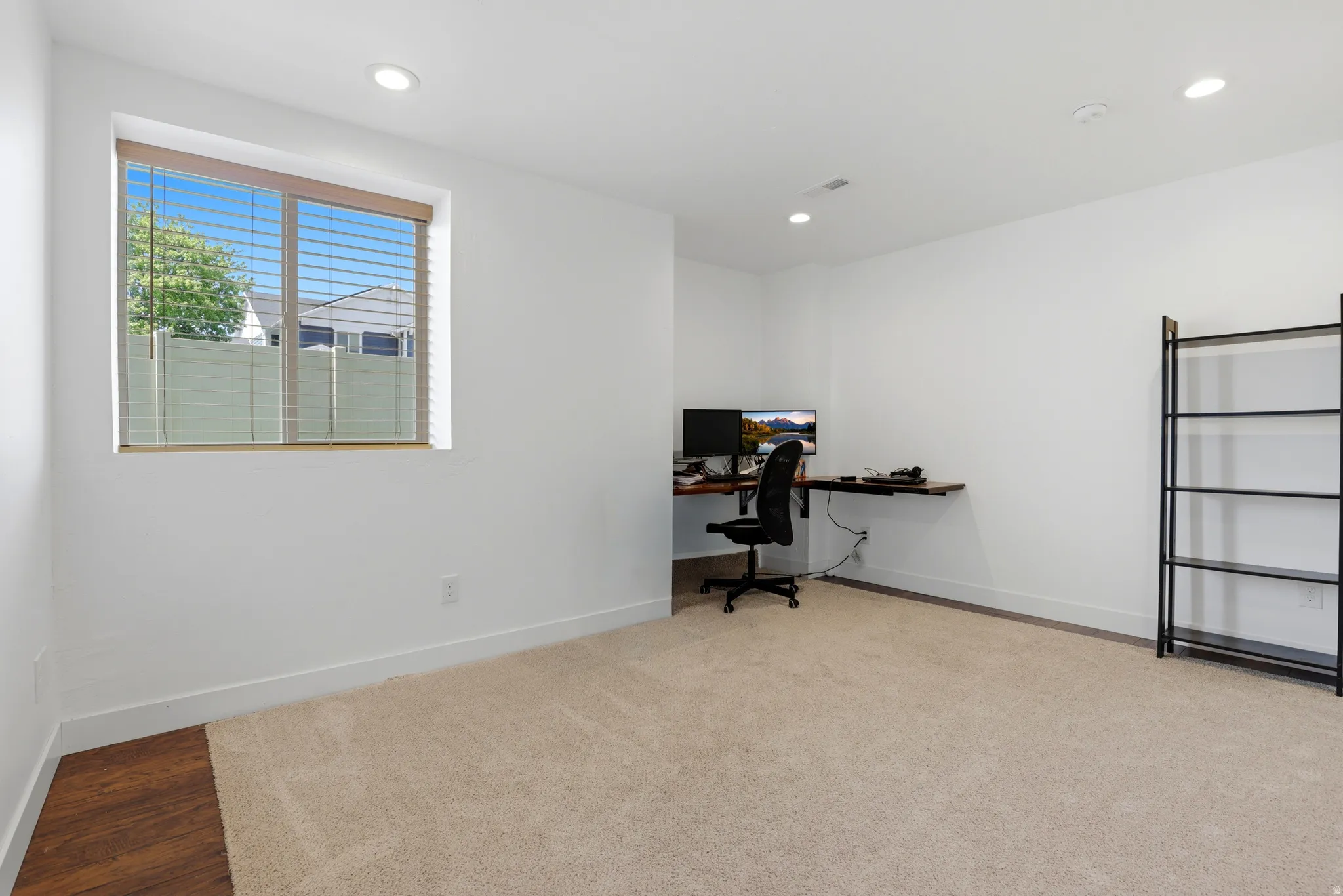 Bright room featuring recessed lighting, a window with horizontal blinds, neutral-toned carpet, white baseboards, and a dedicated corner with wood-finish flooring