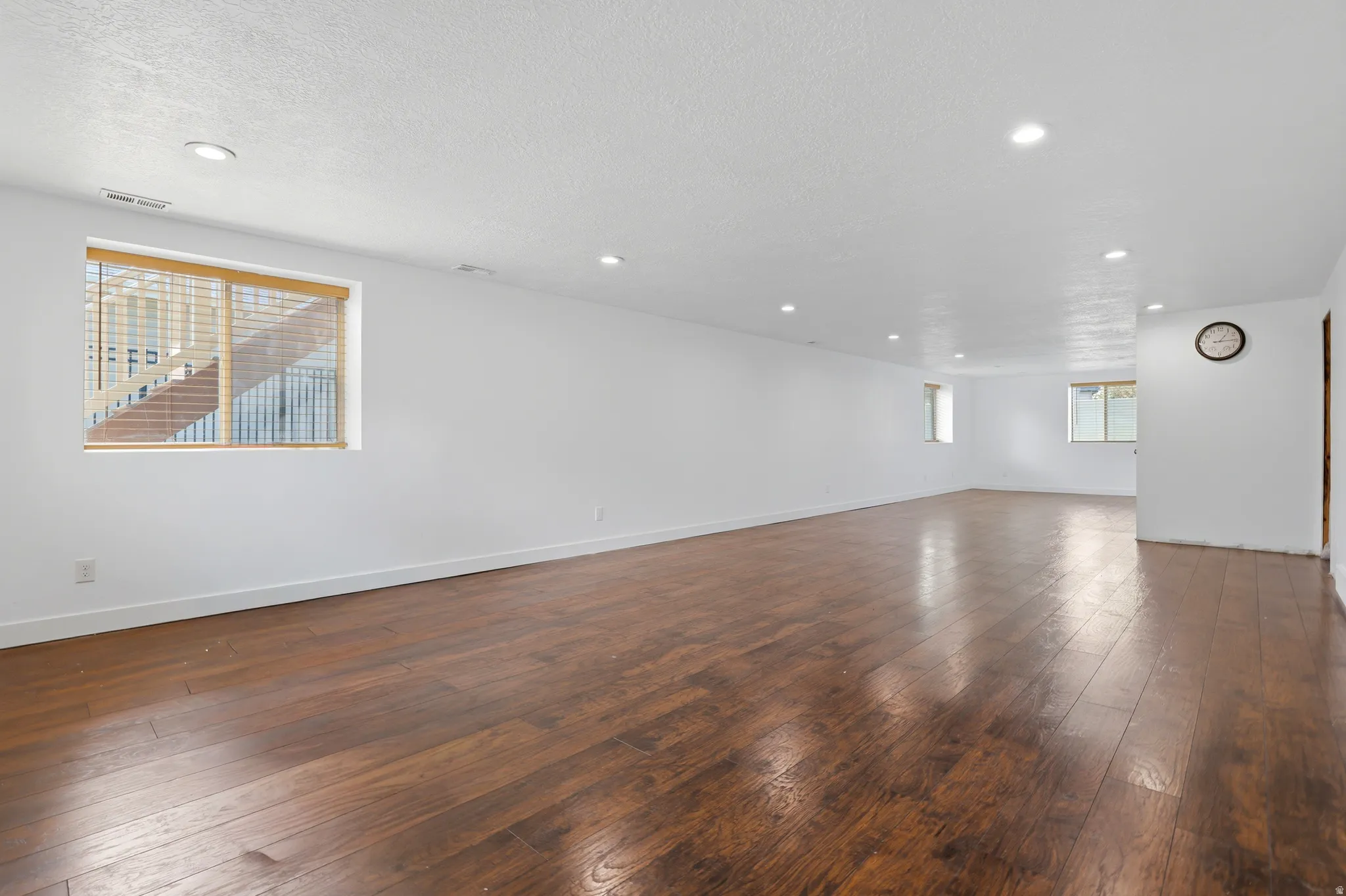 Expansive room featuring rich wood-finish flooring, recessed lighting, multiple windows with blinds, and white painted walls