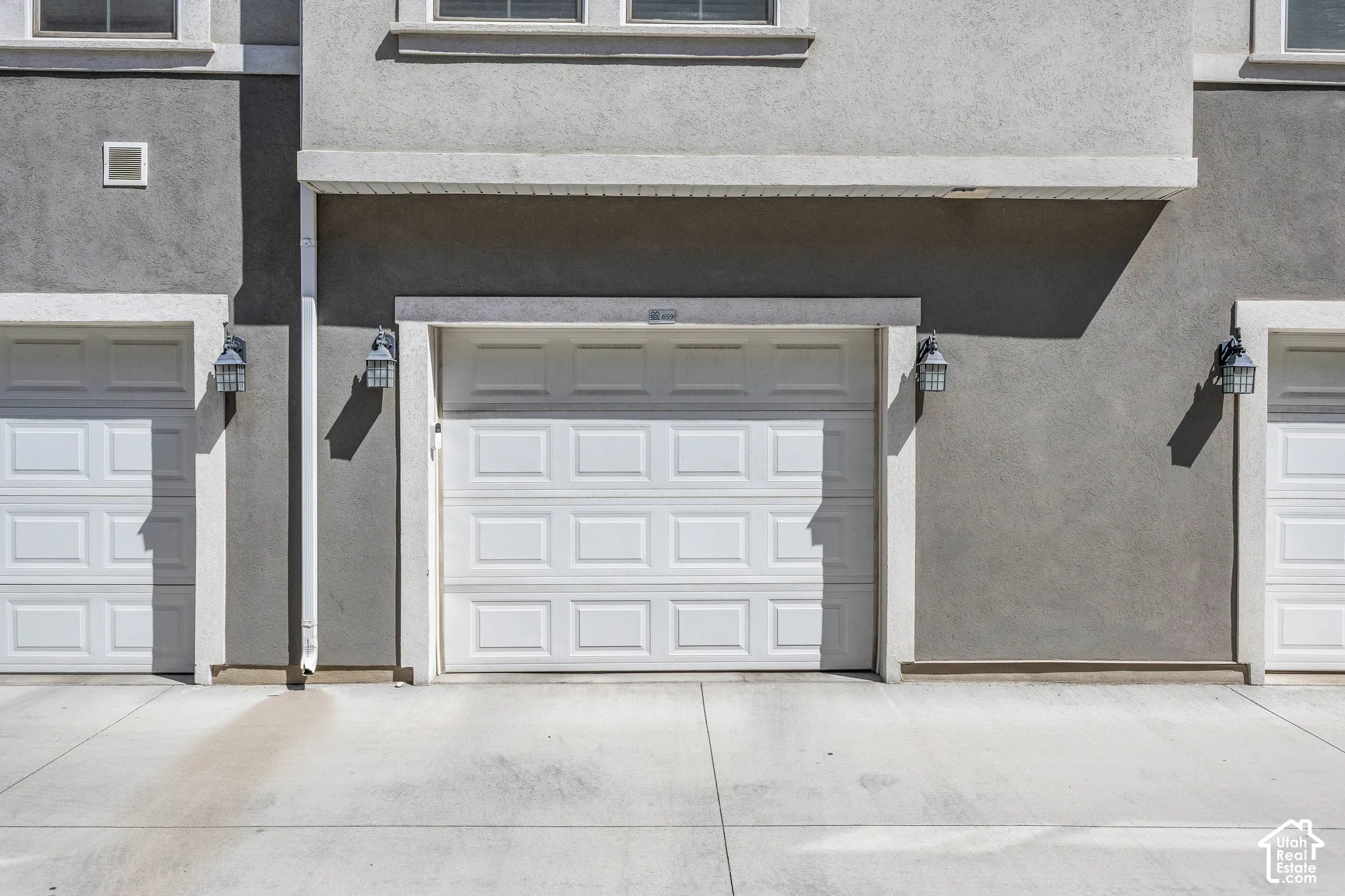 Stucco exterior featuring multiple white paneled garage doors, decorative outdoor lighting fixtures, and a concrete driveway