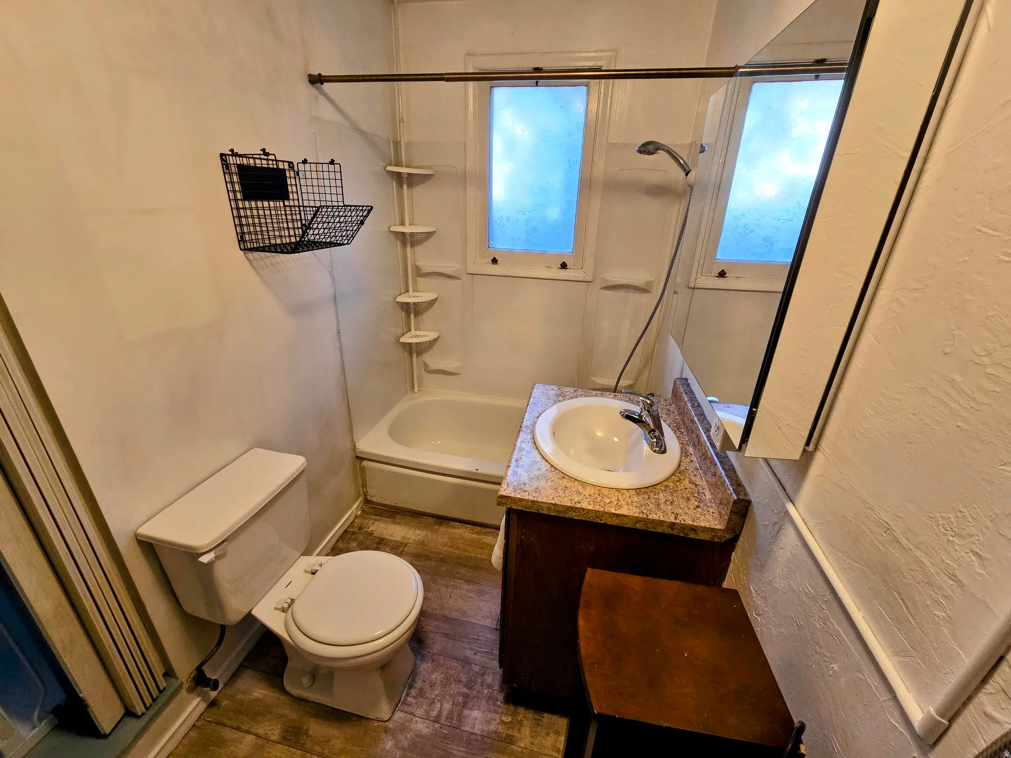 Bathroom featuring wood-finish flooring, a white vanity with an integrated sink and chrome faucet, and a shower-over-tub combination with wall-mounted shelving