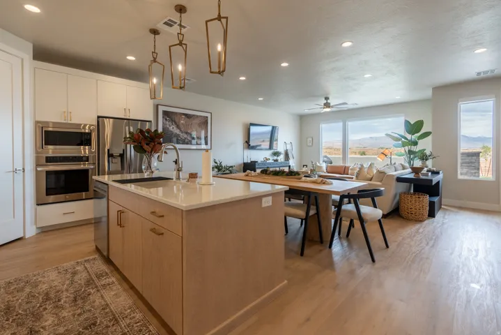 Open-concept interior featuring light wood-finish flooring, a kitchen island with stone countertop, stainless steel appliances, white cabinetry, and large windows revealing a mountain landscape