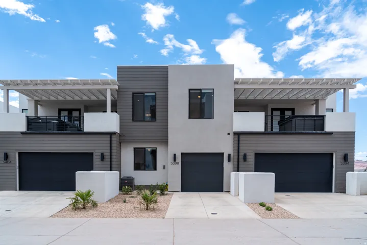 Modern multi-level townhome featuring a stucco and siding exterior, integrated single-car garages, a second-story balcony with metal railings, and a pergola overhead
