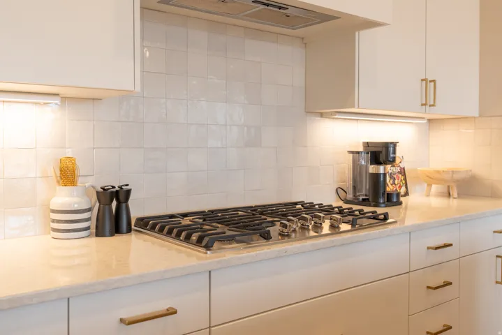 Kitchen featuring a built-in gas cooktop, light-toned countertops, white subway tile backsplash, white cabinetry with gold-toned hardware, and under-cabinet lighting