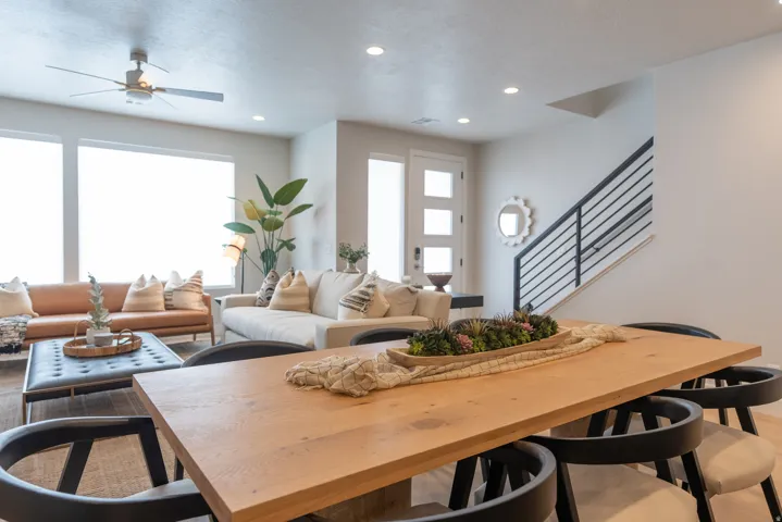 Open-concept living area featuring a light wood-finish dining table, recessed lighting, expansive windows, a modern ceiling fan, and a black metal staircase railing