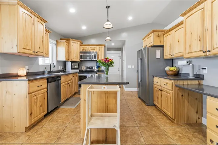 Spacious kitchen featuring light wood cabinetry, dark countertops, a large center island, stainless steel appliances, and tiled flooring