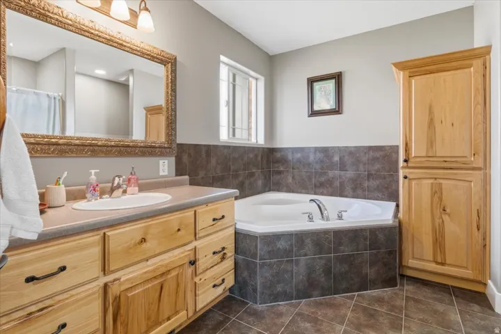 Bathroom featuring a corner soaking tub with tile surround, wood-finish vanity with an integrated sink, framed mirror, and a tall wood-finish storage cabinet