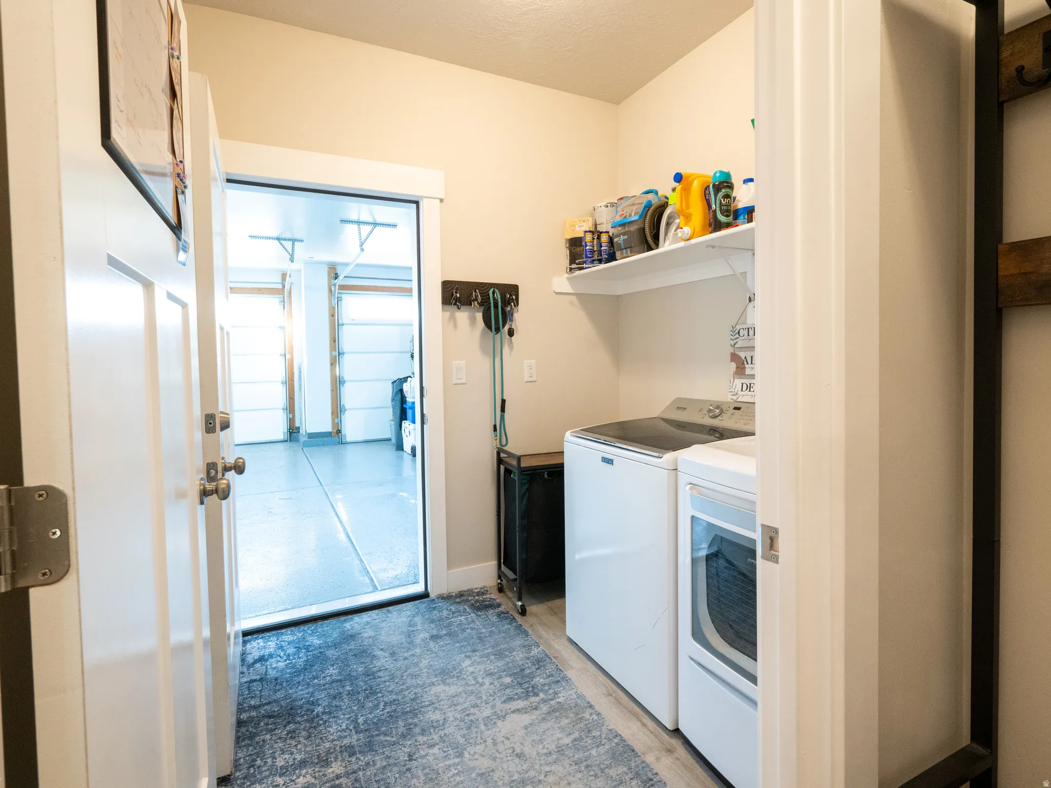 Dedicated laundry area featuring a white floating shelf, light wood-finish flooring, and a doorway opening to a garage with two overhead doors