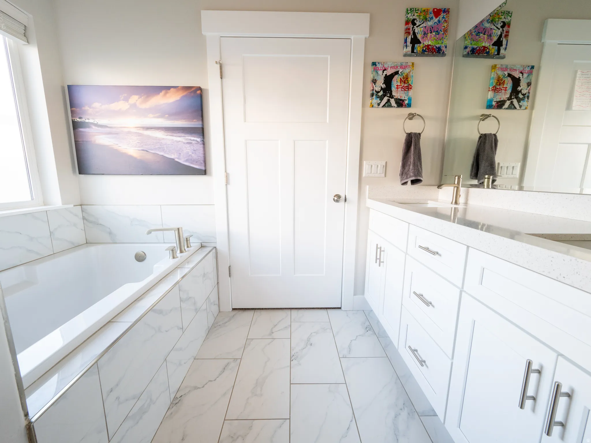 Contemporary bathroom featuring a built-in soaking tub with marble-look tile surround, a spacious double vanity with white cabinetry and light-toned countertops, and marble-look tile flooring
