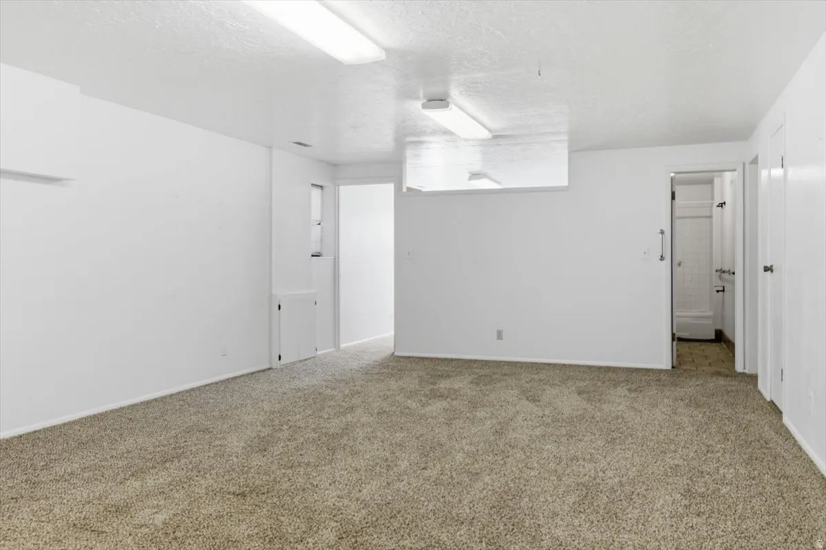 Carpeted room featuring white walls, overhead fluorescent lighting, and a doorway leading to a tiled bathroom with a shower-tub combination