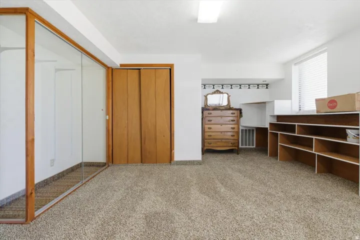 Carpeted interior space featuring a large mirrored closet, wood-finish bi-fold closet doors, built-in shelving, and a window with blinds