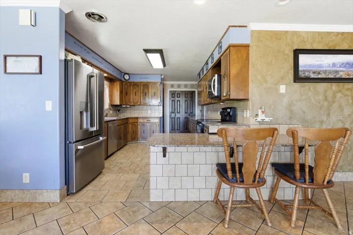 Kitchen featuring tile flooring, stainless steel refrigerator, wood cabinetry, stone countertops, and a tiled breakfast bar