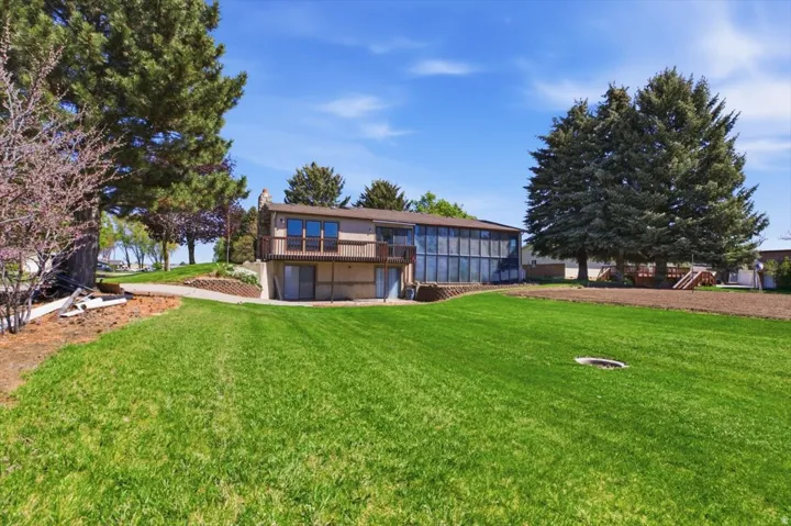 Expansive green lawn with mature evergreen trees and a multi-level residence featuring an upper-level balcony and a lower-level sunroom with extensive window-wall construction