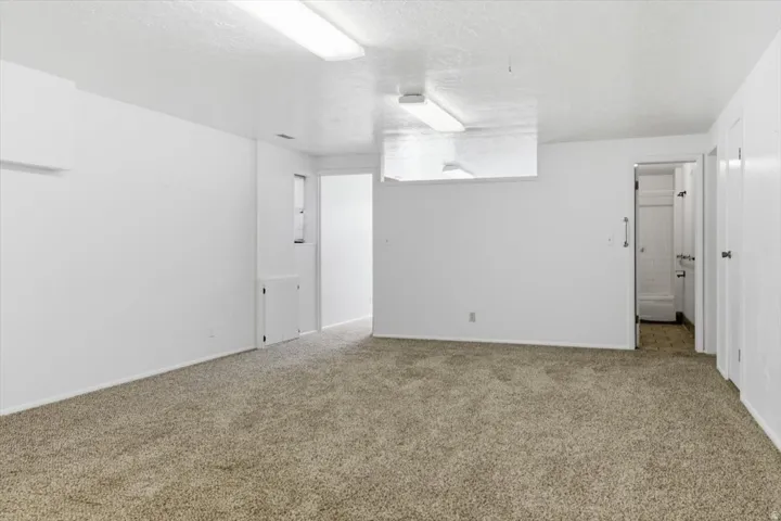 Carpeted room featuring white walls, overhead fluorescent lighting, and a doorway leading to a tiled bathroom with a shower-tub combination