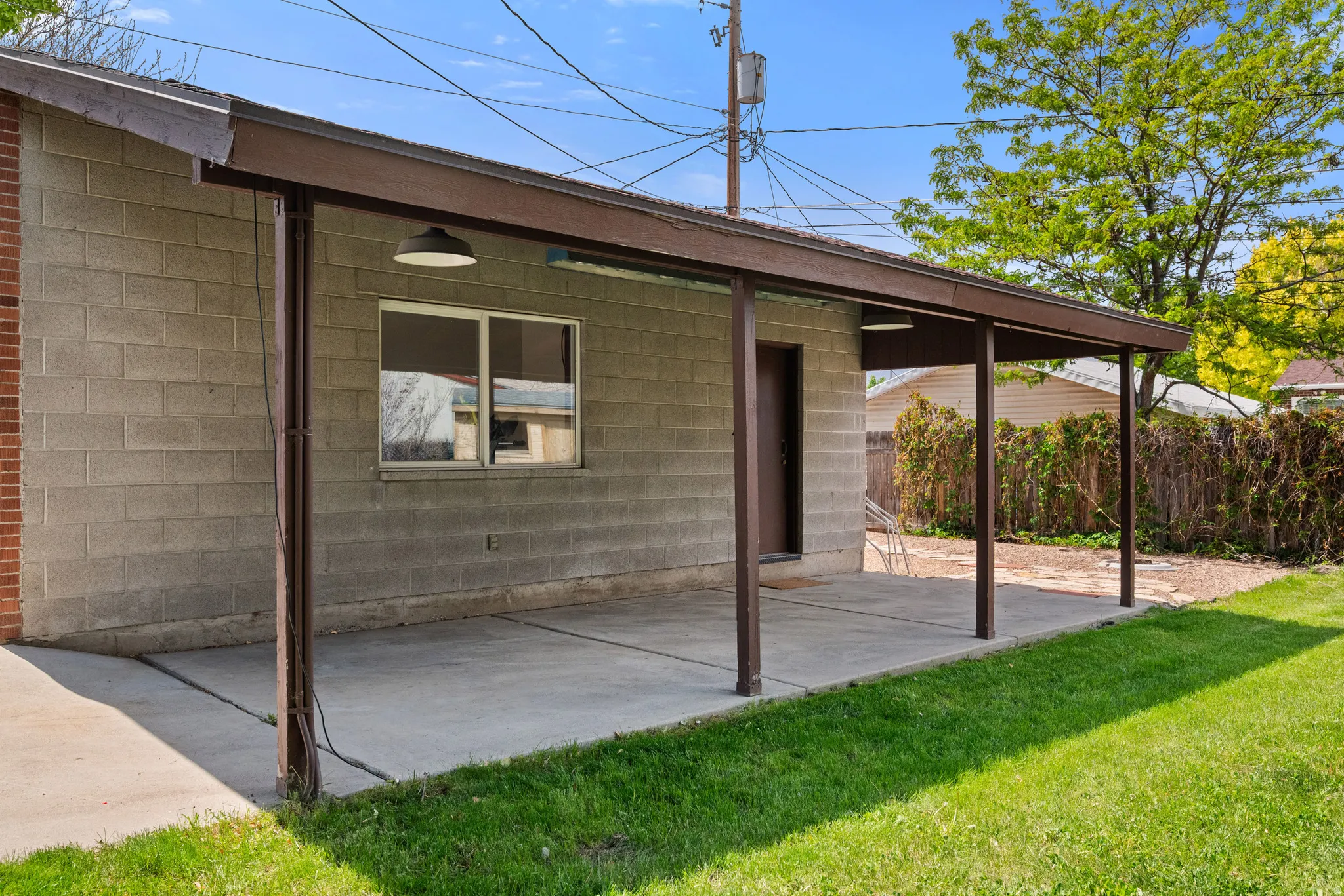 Spacious concrete block exterior featuring a covered patio with a brown frame, a window with exterior lighting, and a brown entry door