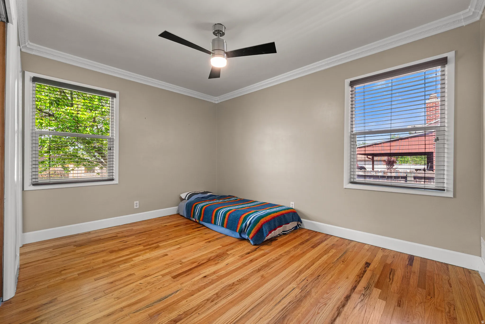 Room featuring wood-finish flooring, two windows with blinds, a modern ceiling fan, crown molding, and baseboards