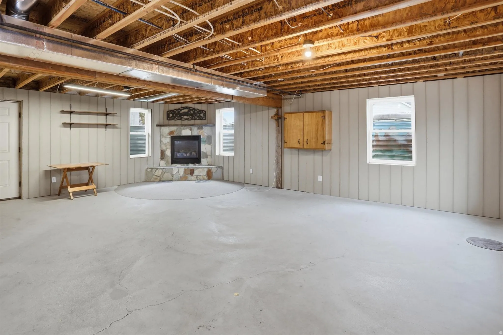 Spacious unfinished basement featuring a stone fireplace, exposed ceiling joists, light-colored panel walls, multiple windows, and a concrete floor