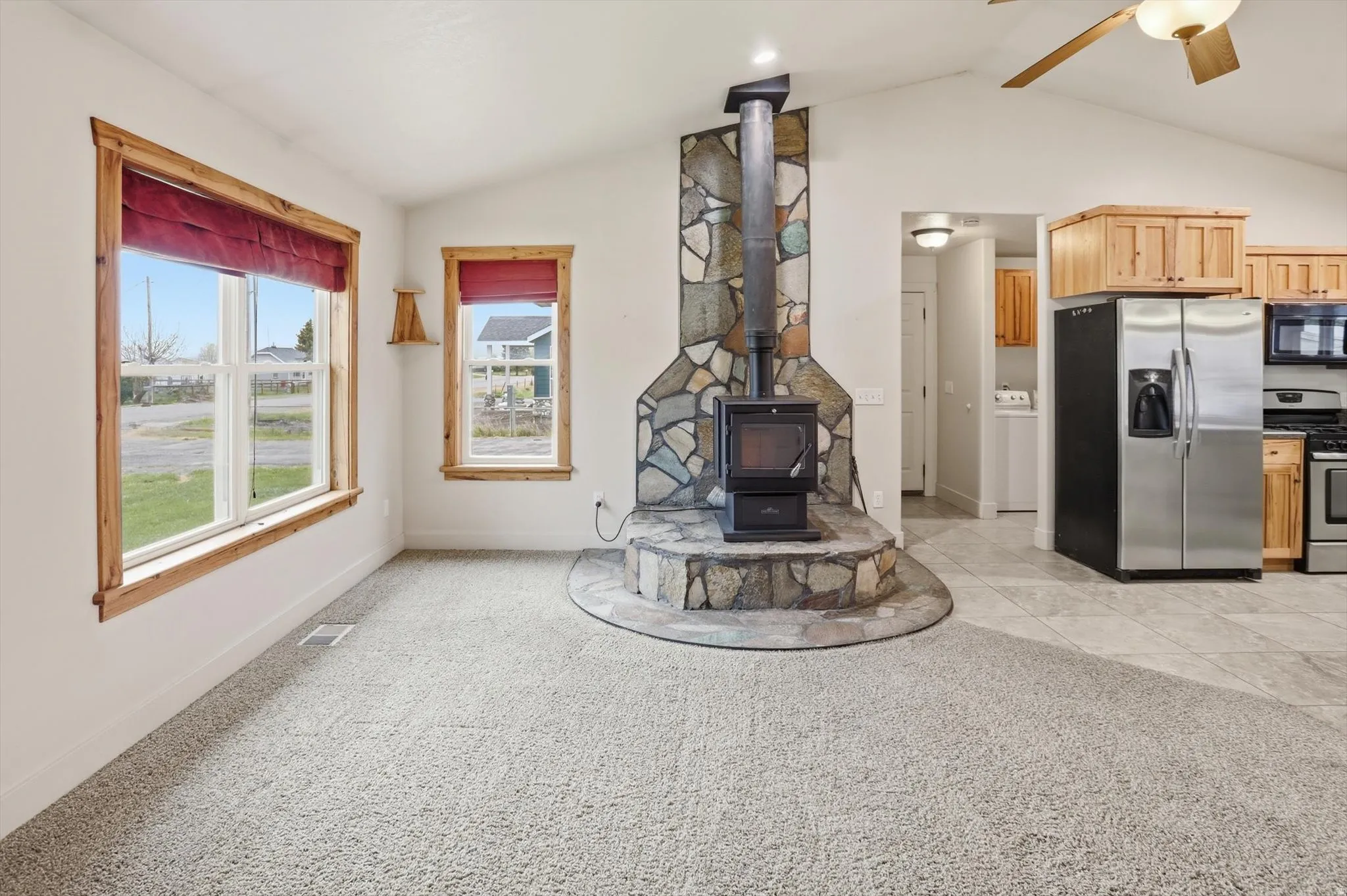 Vaulted ceiling living space featuring a stone-clad wood-burning stove with a raised hearth, light-toned carpet, and natural wood window trim