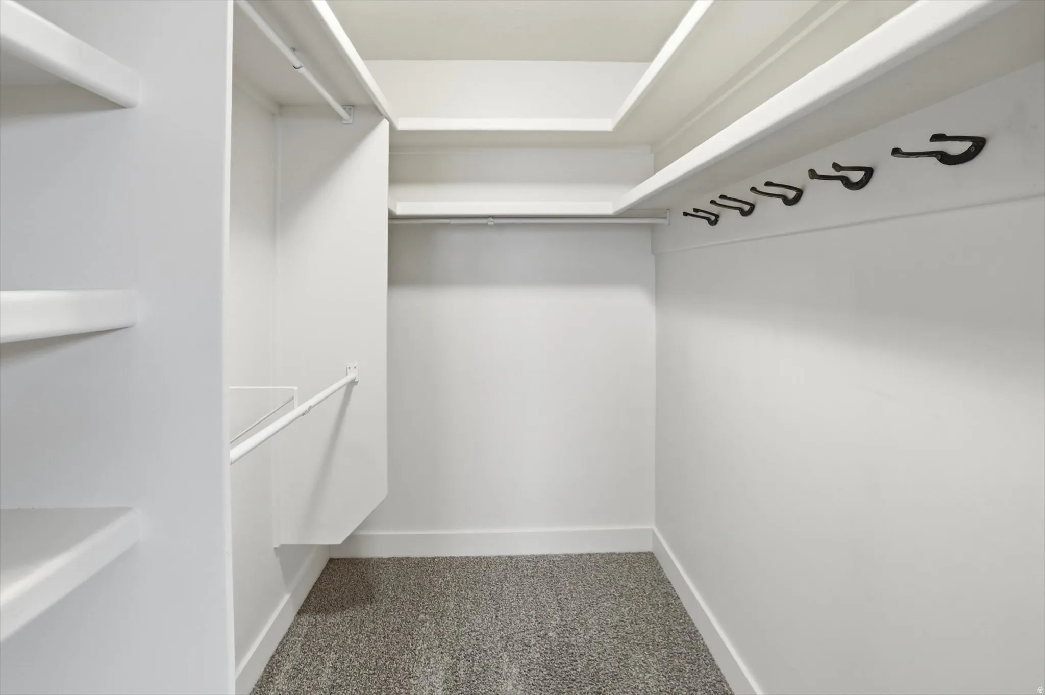 Walk-in closet featuring white built-in shelving, white walls, black wall hooks, and gray speckled flooring