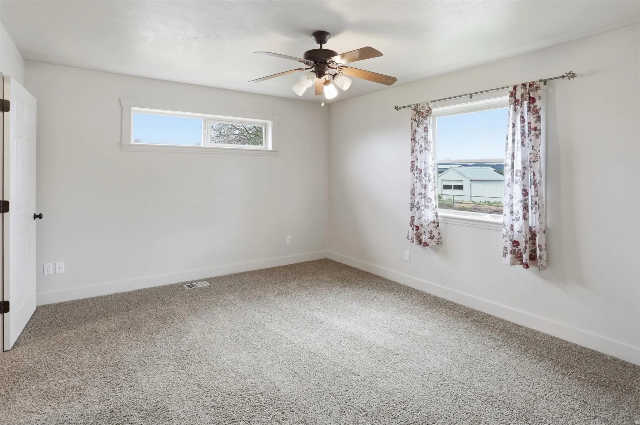 Carpeted room featuring a ceiling fan with light fixture, two windows providing natural illumination, white trim, and a white interior door