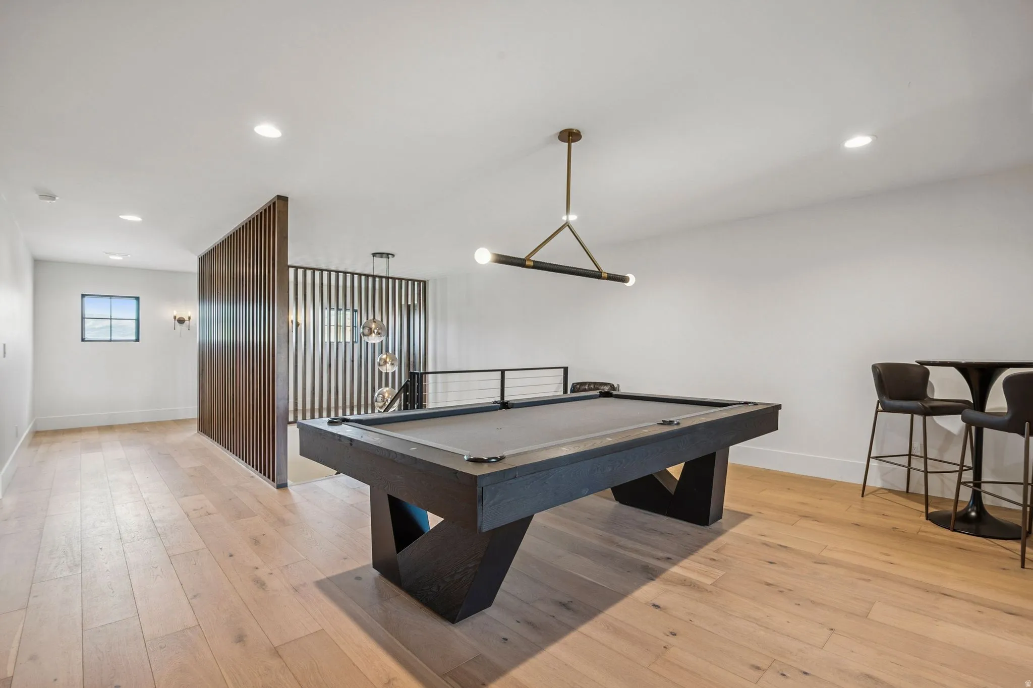 Spacious entertainment area featuring light hardwood flooring, a modern linear chandelier, recessed lighting, a decorative wood slat partition, and black metal stair railing