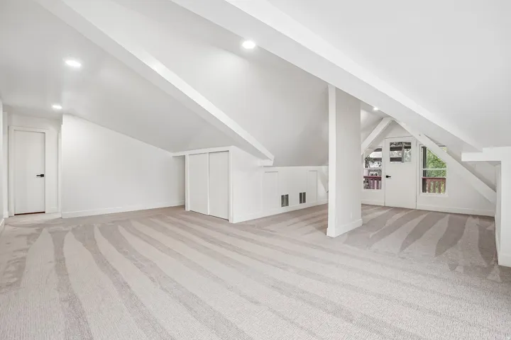 Spacious carpeted room featuring a sloped ceiling with exposed beams, recessed lighting, and a white interior door