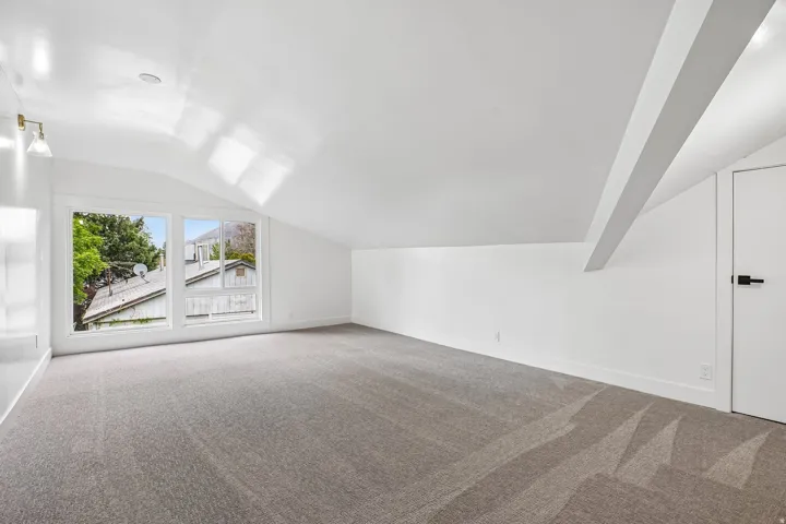 Spacious room featuring vaulted ceilings, a large multi-pane window, flush-mounted ceiling lighting, wall-mounted sconce lighting, and soft grey carpeting