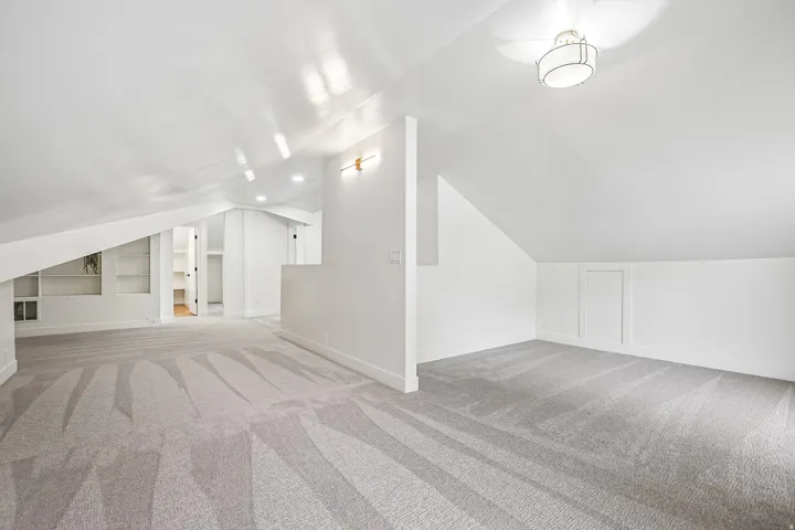 Spacious loft area featuring sloped ceilings, recessed lighting, wall-mounted sconces, built-in shelving, and light gray patterned carpeting