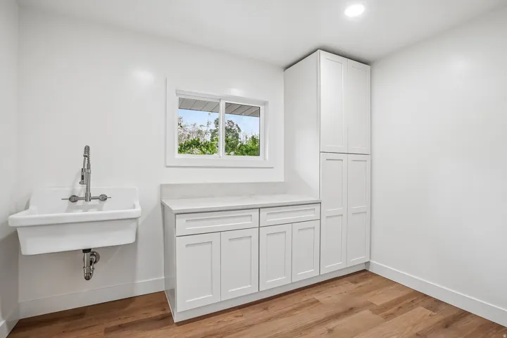Utility room featuring a wall-mounted laundry sink with a pull-down faucet, white shaker cabinetry, light-toned countertop, and wood-finish flooring