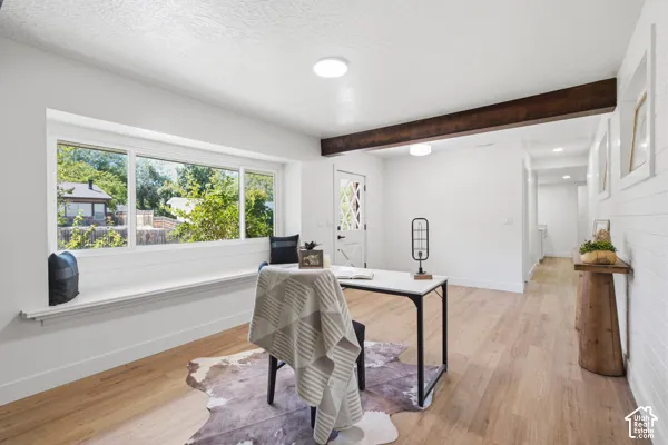 Bright home office featuring expansive multi-pane windows with natural light, warm wood-finish flooring, exposed ceiling beam detail, and a minimalist workspace with clean white walls and recessed lighting