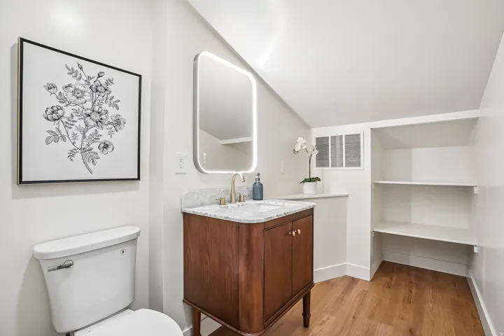 Bathroom featuring a wood-finish vanity with a stone countertop, an illuminated mirror, a white toilet, built-in shelving, and wood-finish flooring