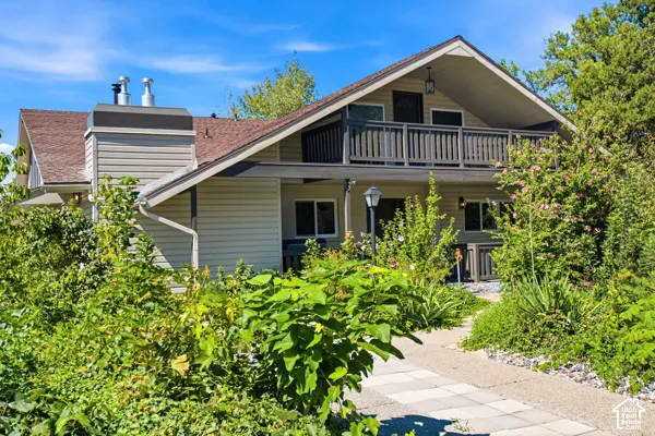Two-story exterior with siding, a second-floor balcony featuring wood railing, a shingled roof, a paved walkway, and a prominent chimney
