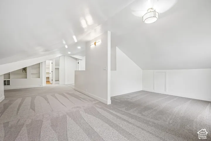 Spacious loft area featuring sloped ceilings, recessed lighting, wall-mounted sconces, built-in shelving, and light gray patterned carpeting