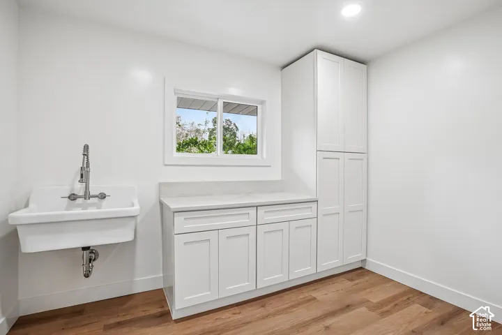 Utility room featuring a wall-mounted laundry sink with a pull-down faucet, white shaker cabinetry, light-toned countertop, and wood-finish flooring
