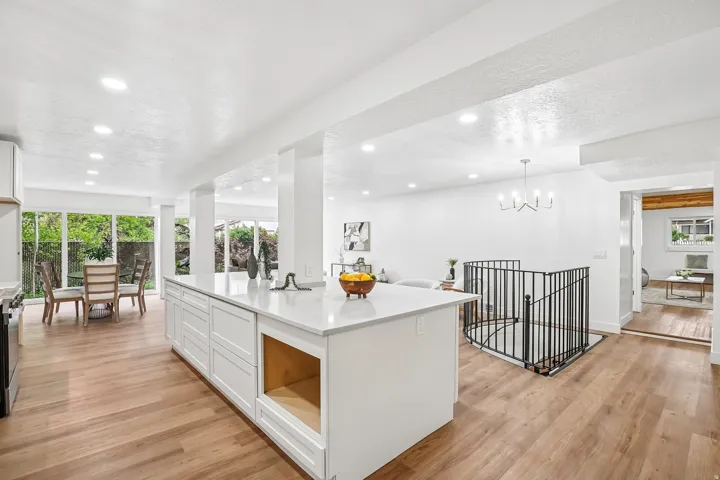 Expansive kitchen island with a light-toned countertop, wood-finish flooring, recessed lighting, white cabinetry, and a black metal spindle staircase
