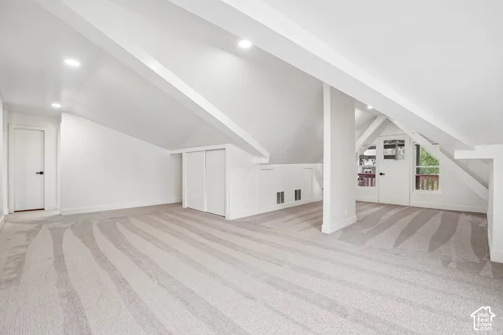 Spacious carpeted room featuring a sloped ceiling with exposed beams, recessed lighting, and a white interior door