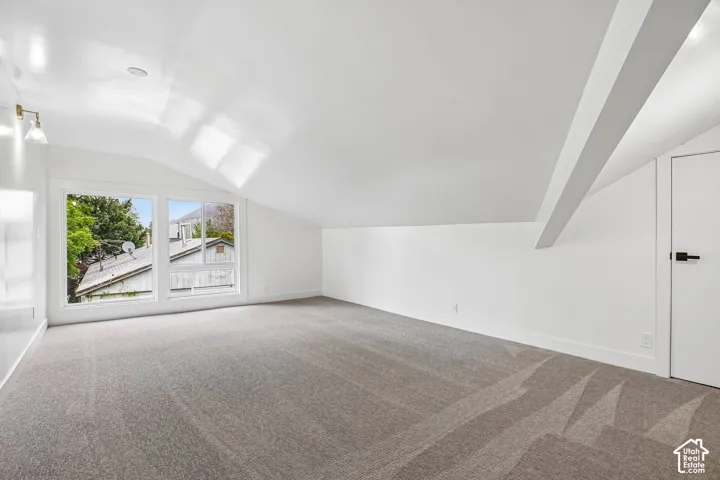 Spacious room featuring vaulted ceilings, a large multi-pane window, flush-mounted ceiling lighting, wall-mounted sconce lighting, and soft grey carpeting