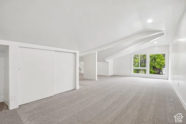 Spacious room featuring light gray carpeting, recessed lighting, white walls, angled ceilings with structural beams, and a window with multiple panes