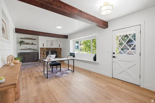 Hardwood-style flooring, exposed wood ceiling beams, stone-clad fireplace with built-in shelving, window with integrated seating, and a decorative paneled entry door
