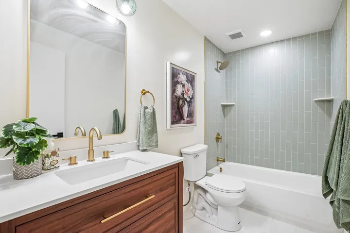 Bathroom featuring a wood-finish vanity with a white countertop and rectangular undermount sink, brushed gold hardware and faucet, and a shower-tub combination with light green subway tile surround and integrated shelving