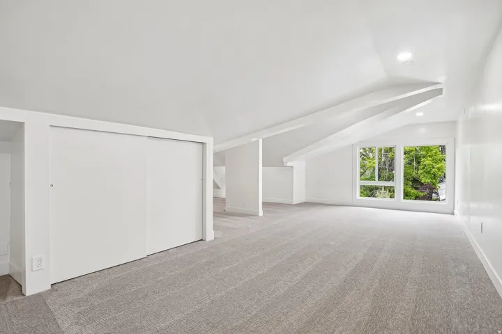 Spacious room featuring light gray carpeting, recessed lighting, white walls, angled ceilings with structural beams, and a window with multiple panes